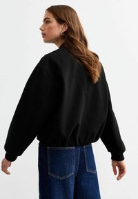 Felpa nera con vestibilità over-size, polsini e orlo a costine. Indossata con jeans blu a vita alta, mostrando una texture liscia e linee di design pulite.
