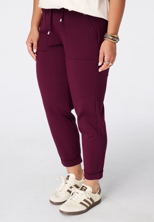 Vrouw met opgerolde bordeauxrode broek, witte sneakers met bruine strepen en gouden en zilveren armbanden om haar rechterpols.