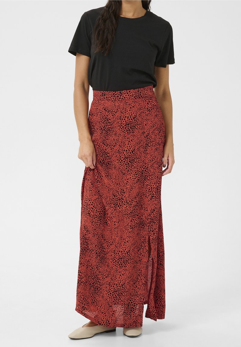 Femme portant un top noir à manches courtes avec une jupe maxi rouge taille haute présentant des taches abstraites noires et une fente latérale, associée à des ballerines beiges.