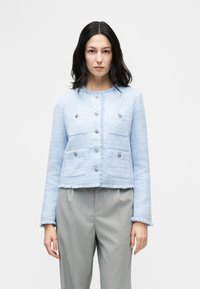 Γυναίκα με σκούρα μαλλιά που φοράει ένα γαλάζιο υφασμάτινο σακάκι με διακοσμητικά κουμπιά και γκρι παντελόνι με ρίγες, στέκεται μπροστά σε απλό φόντο.