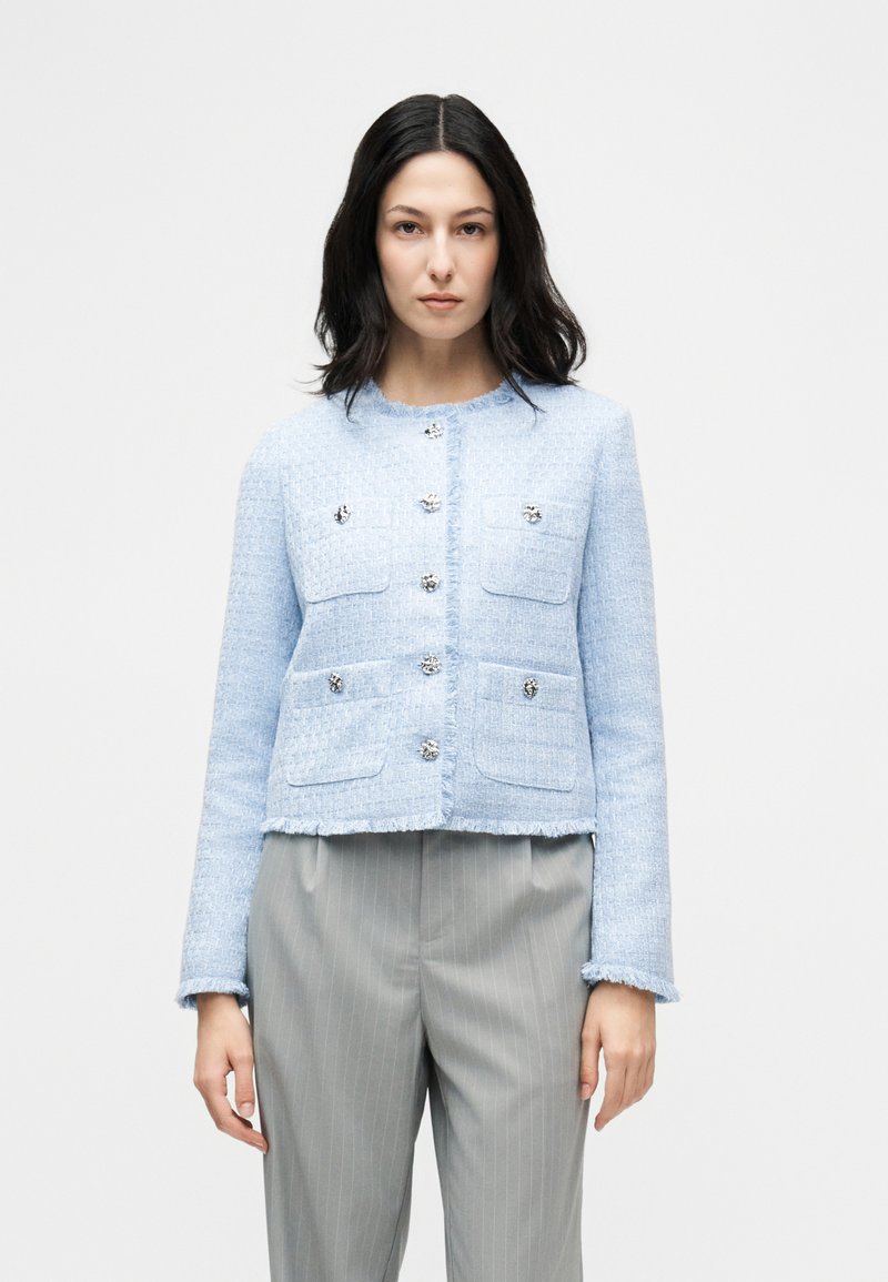 Γυναίκα με σκούρα μαλλιά που φοράει ένα γαλάζιο υφασμάτινο σακάκι με διακοσμητικά κουμπιά και γκρι παντελόνι με ρίγες, στέκεται μπροστά σε απλό φόντο.