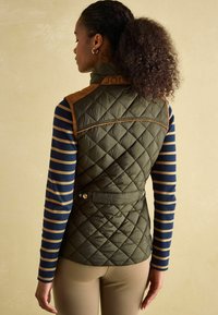 Prešívaná oliva vestu s hnedými ramennými akcentmi, vysokým golierom a detailmi v oblasti pásu, nosená cez dlhý rukáv tričko s modro-bežovými pruhmi.