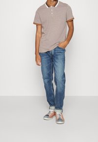 Camisa polo marrón con cuello a rayas blancas y marrones, combinada con jeans azules enrollados en los puños y zapatillas de deporte marrones claras con punteras grises.
