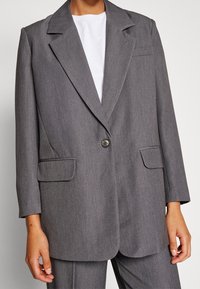 Blazer grigio su misura con chiusura a un bottone, rever a toppa, due tasche frontali e una texture liscia. Indossato sopra un top bianco.