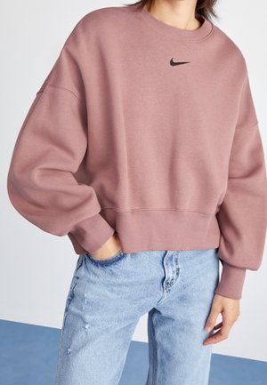 Personne portant un sweat-shirt Nike rose poudré avec un logo noir, associé à un jean taille haute bleu clair, debout avec une main dans la poche.