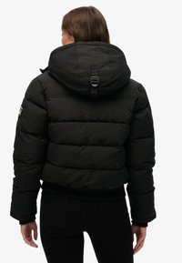 Sort dunjakke med hætte og ribbede manchetter. Har vandrette syninger og et logopatch på ærmet. Materialet virker tekstureret.