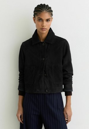 Žena s copmi má na sebe čiernu zapínaciu bundu s prednými vreckami a námornícky modré nohavice s pruhmi, stojaca pri bielej stene.