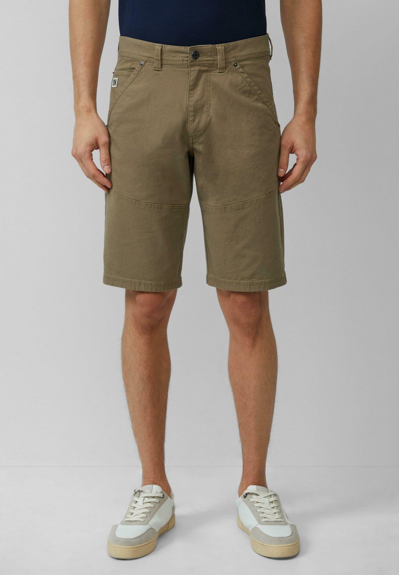 Beige Baumwollshorts mit lockerer Passform, ausgestattet mit einem Reißverschluss, Fronttaschen und einem knielangen Design; kombiniert mit hellen Sneakers.