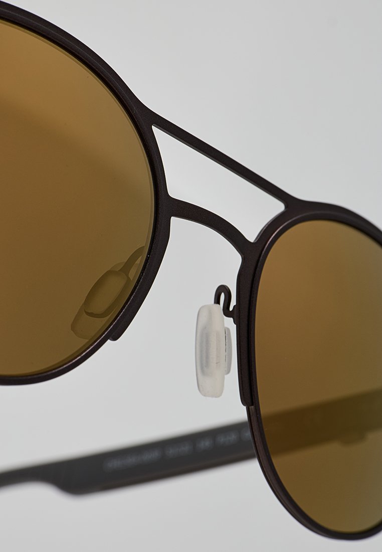 Gafas de sol con tinte marrón y marco de metal negro mate, que presentan un diseño geométrico único y almohadillas nasales ajustables para mayor comodidad.