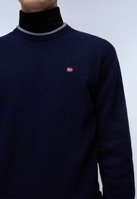Marineblauwe gebreide trui met een hoge kraag, voorzien van een grijze rand en een klein rood-wit geborduurd vlagdetail op de borst.