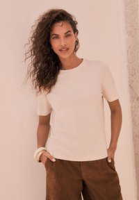 Maglietta a maniche corte beige chiaro con scollo rotondo, abbinata a pantaloni in suede marrone. Presenta una texture liscia e un design sottile.