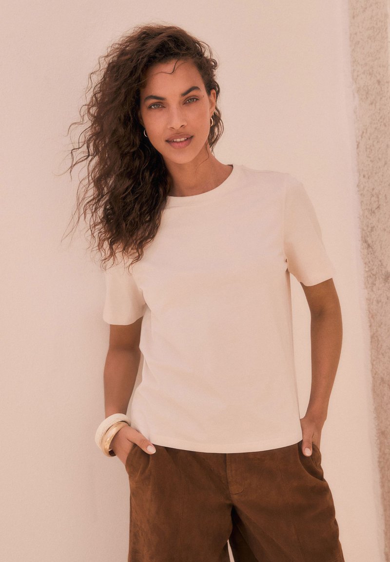 Maglietta a maniche corte beige chiaro con scollo rotondo, abbinata a pantaloni in suede marrone. Presenta una texture liscia e un design sottile.