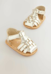 Metallic gold sandals featuring a woven design, open toe, and Velcro strap, set on a natural-colored, flat sole.