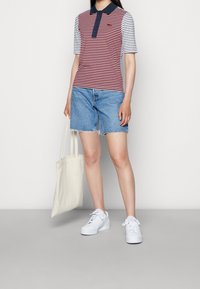 Polo rayé rouge et blanc avec un col bleu marine, associé à un short en jean clair et des baskets blanches, tenant un sac fourre-tout crème.