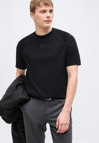 Νέος άνδρας που φοράει μαύρο κοντομάνικο πουλόβερ Porsche Boss και γκρι παντελόνι, κρατώντας ένα μαύρο σακάκι στο αριστερό του χέρι.