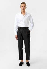 Witte slim-fit shirt, zwarte geplooide broek met hoge taille en knoopaccenten, afgewerkt met zwarte loafers. Glad stoftextuur.