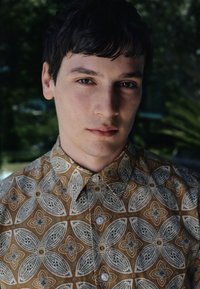 Joven con pelo corto y oscuro que lleva una camisa marrón y blanca con botones y estampado, de pie al aire libre con un fondo borroso de follaje verde.
