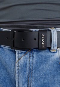 Black leather belt with rectangular buckle; brand logo embossed in white; worn with light blue denim jeans.
