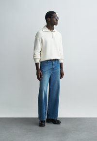 Muž v bílém dlouhém pulovru, modrých džínách s širokými nohavicemi, černých botách, brýlích a perlovém náhrdelníku stojí před jednobarevným světlým pozadím.