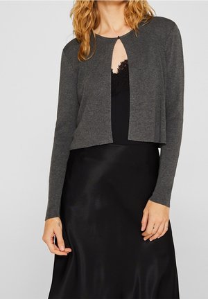 Frau trägt ein dunkelgraues, kurz geschnittenes Strickjäckchen über einem schwarzen Top mit Spitzenbesatz und einem schwarzen Satinhrock, steht vor hellem Hintergrund.