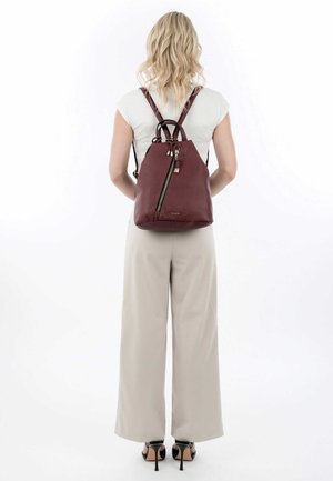 Sac à dos en cuir bordeaux avec une forme effilée, des bretelles doubles et une fermeture éclair bien visible. Surface texturée avec des accents métalliques.