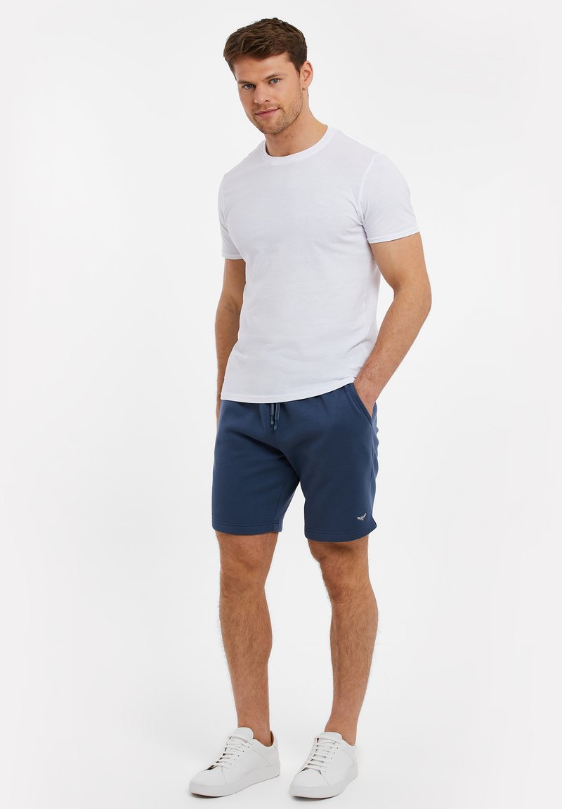 T-shirt en coton blanc associé à un short marine avec une taille élastique et des poches latérales ; le modèle porte des baskets blanches.