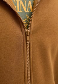 Közeli kép egy részben becipzárazott barna kabátról, amely alatt egy zöld póló látható sárga szöveggel és grafikával.