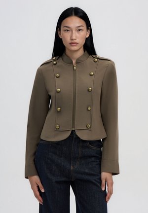 Donna con lunghi capelli neri che indossa una giacca corta verde oliva con bottoni dorati e jeans blu scuro a vita alta, in piedi davanti a uno sfondo semplice.