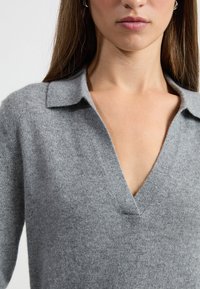 Szary sweter dzianinowy z dekoltem w kształcie litery V i krótkim kołnierzykiem. Posiada miękką strukturę oraz subtelny, równomierny wzór dzianiny.