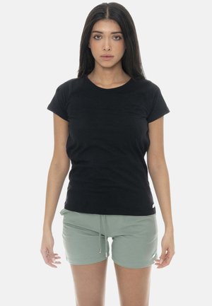 Femme aux longs cheveux foncés portant un t-shirt noir cintré à manches courtes et un short vert clair à cordon, debout devant un fond uni.