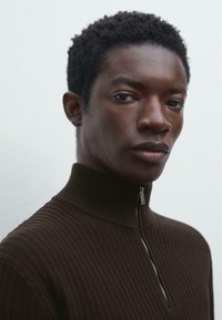 Brauner, gerippter Pullover mit hohem Kragen und Frontreißverschluss. Glatte Textur, enganliegendes Design mit subtilen horizontalen Linien.