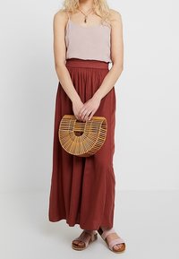 Rosa spaghetti strap topp, rostfärgad maxikjol, rosa sandaler, håller en rund bambuväska. Enkel design, avslappnad passform, lätt tyg.