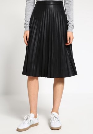 Person iført et sort plisseret midi-nederdel, lysegrå bluse med lange ærmer og hvide sneakers med gummisåler på en lys baggrund.