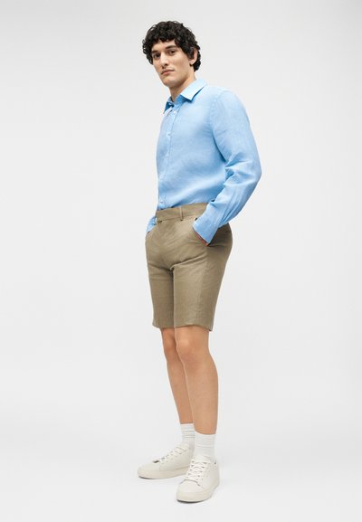 Άνδρας με ανοιχτό μπλε πουκάμισο μακρύ μανίκι, μπεζ σορτς, λευκές κάλτσες και λευκά αθλητικά παπούτσια, στέκεται με τα χέρια στις τσέπες σε μονόχρωμο φόντο.