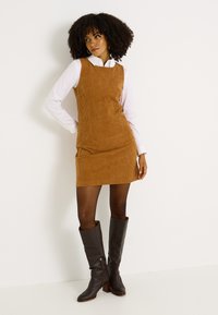 Vestido de pana en marrón con una forma ajustada, combinado con botas de cuero marrón hasta la rodilla, acentuando un look en capas con una camiseta blanca debajo.