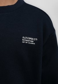 Maglione in cotone blu navy con collo a giro, caratterizzato da testo bianco che recita "ASSEMBLED PETROLIND AW'89 ALASKA" sul petto.