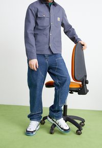 Una persona in giacca da lavoro blu navy con diverse tasche, jeans blu strappati e sneaker bianche si trova accanto a una sedia da ufficio con accenti arancioni.