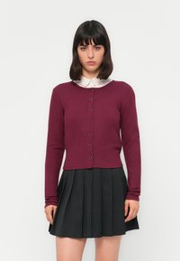 Femme aux cheveux sombres et lisses portant un cardigan bordeaux boutonné sur une chemise blanche à col et une jupe plissée noire, se tenant devant un fond blanc.