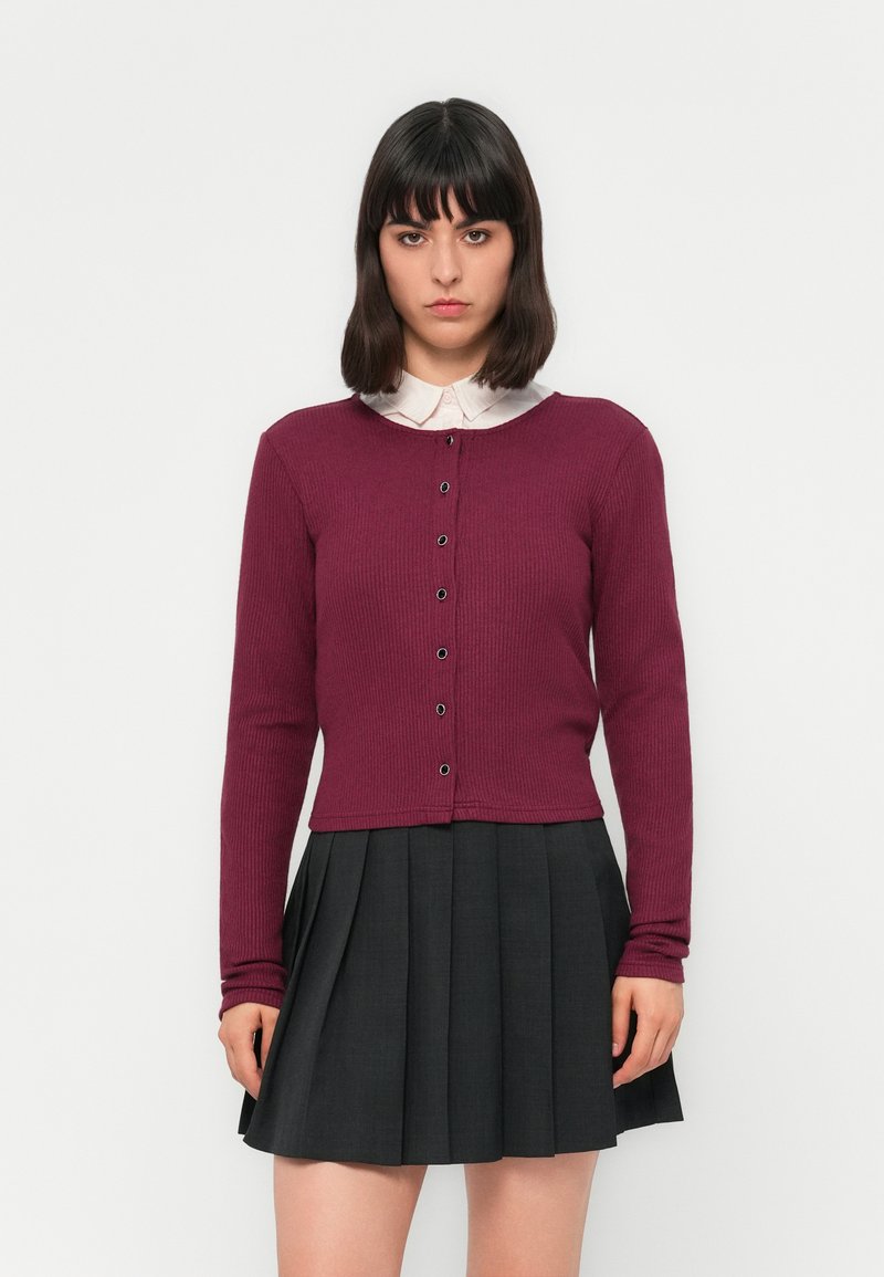 Femme aux cheveux sombres et lisses portant un cardigan bordeaux boutonné sur une chemise blanche à col et une jupe plissée noire, se tenant devant un fond blanc.