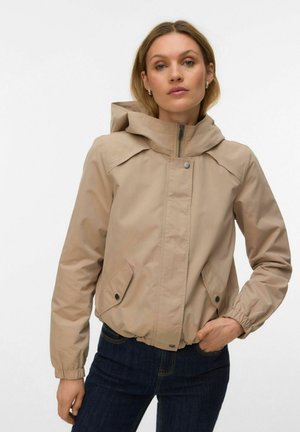 Kobieta ubrana w beżową kurtkę z kapturem zapinaną na zamek z kieszeniami na guziki, w połączeniu z ciemnoniebieskimi dżinsami, stoi na jasnym, jednolitym tle.