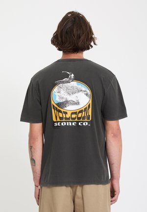Personne aux cheveux bruns portant un t-shirt noir Volcom Stone Co. avec un motif de surf et du texte au dos, associé à un pantalon beige.