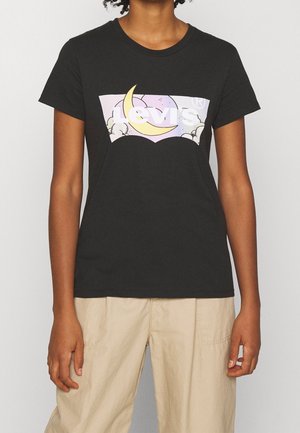 Personne portant un t-shirt noir Levi's avec un graphique de ciel nocturne pastel, croissant de lune et nuages, associé à un pantalon beige.