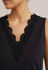 Črna brezrokavnica s V-izrezom in zapletenimi podrobnostmi iz črnega čipke ter gladkim tekstilnim videzom.
