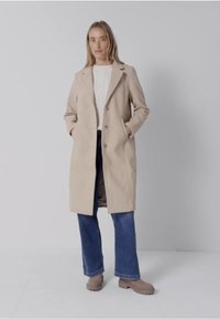 Žena stojící s rukama v kapsách, oblečená v béžovém dlouhém kabátu, bílém svetru, modrých džínách a béžových kotníkových botách na jednolitém pozadí.