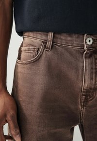 Blugi din denim maro cu o textură netedă și un luciu subtil, cu buzunare frontale, un buton argintiu și detalii de cusătură contrastante.