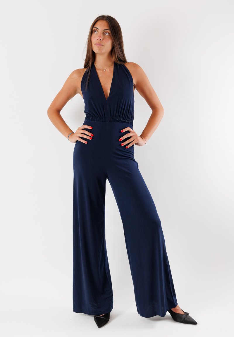 Tuta blu navy con collo a halter, scollatura profonda a V, vita arricciata, pantaloni larghi e superficie liscia. Indossato con tacchi a punta neri.