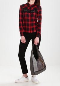 Chemise boutonnée à carreaux rouge et noir avec détail en dentelle, pantalon noir et sac à épaule en cuir gris avec des accents cousus. Baskets blanches.