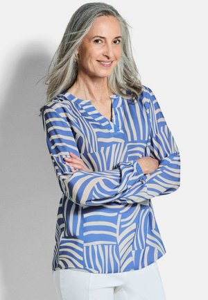 Glimlachende vrouw met lang grijs haar, gekleed in een blauw-beige blouse met abstracte strepen en een witte broek, met gekruiste armen.