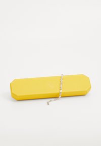 Caixa de joias amarela de forma octogonal com uma superfície texturizada, contendo uma pulseira de prata com pedras transparentes em corte oval dispostas em um padrão linear.