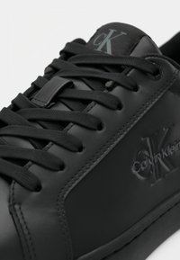 Zwarte leren sneakers met zwarte veters, met het reliëf Calvin Klein-logo aan de zijkant en "cK" gedrukt op de tong.